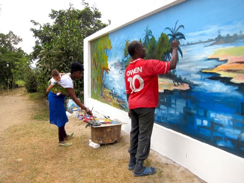 Portant son fils Quentin sur le dos, Rhode Makoumbou réalise la fresque murale «Le village de pêcheurs»