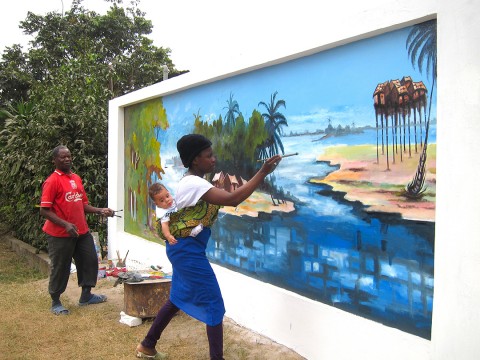 Rhode Makoumbou portant son fils Quentin et réalisant en même temps la fresque murale «Le village de pêcheurs»